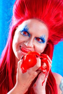 A woman wearing a large red wig with red contact lenses an blue eye make up shaped like flames, while eating a bleeding apple