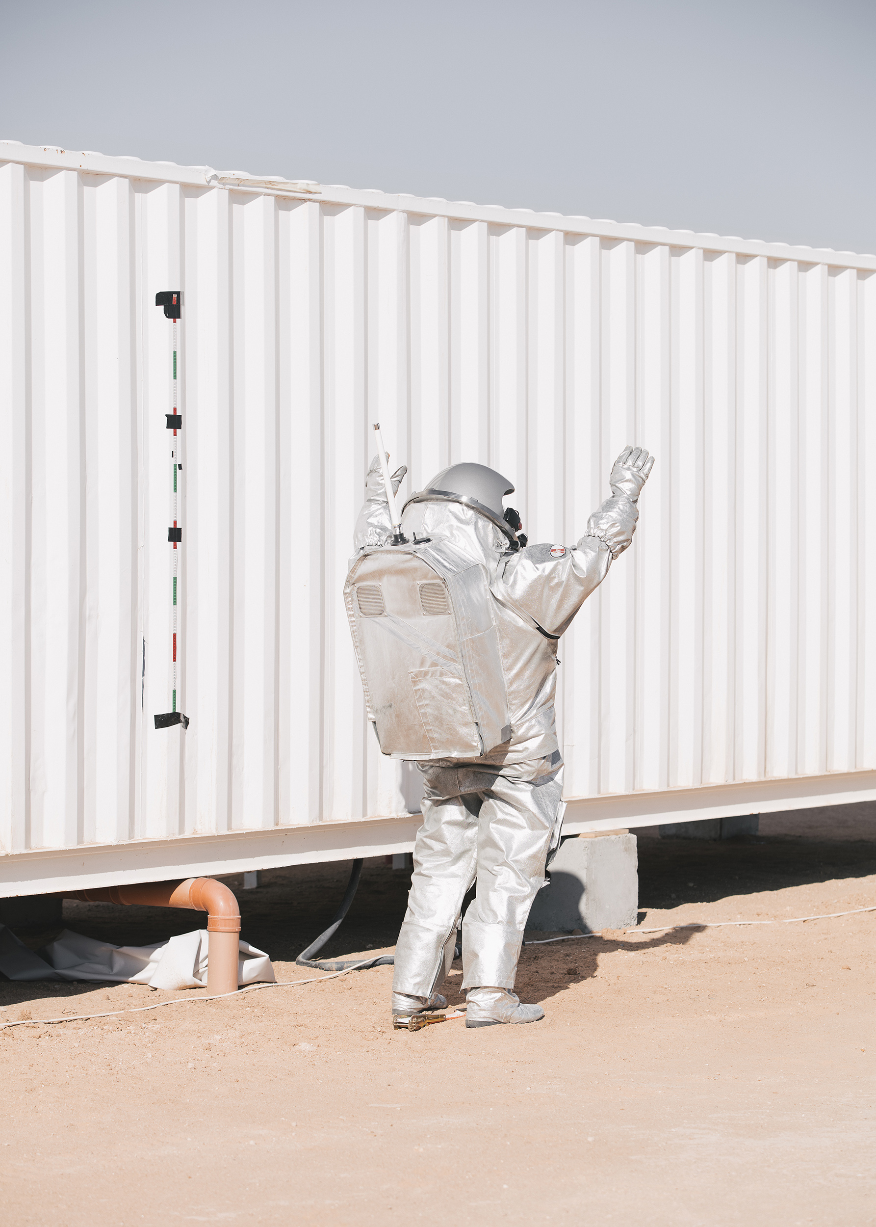 Ein Mensch steht in einem silbernen Raumanzug in der Wüste neben einem Container und streckt die Arme
