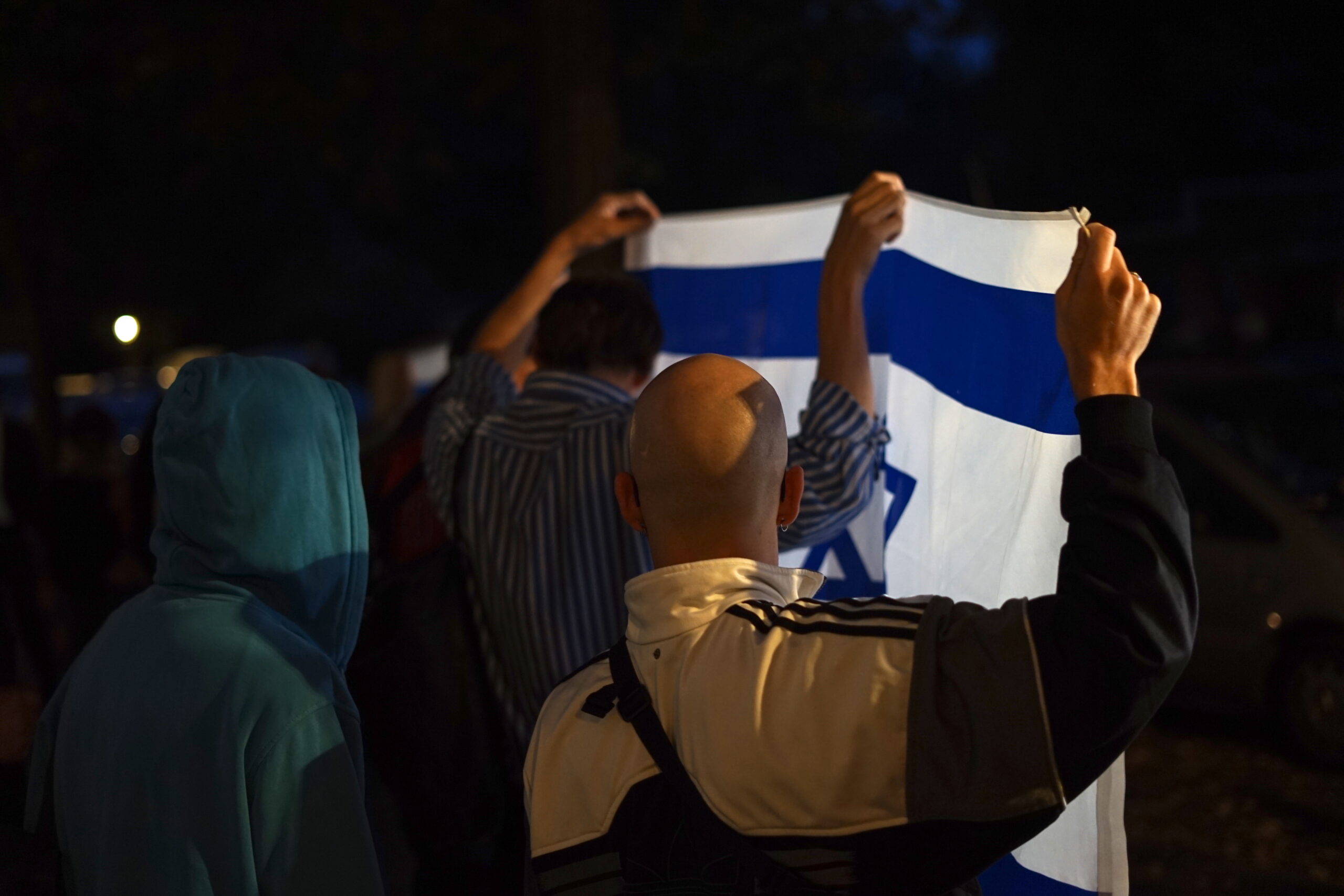 Zwei Männer, den Rücken zur Kamera, halten im Dunklen eine Israel-Fahne hoch