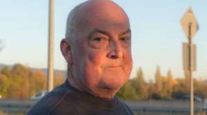 Ein Mann mit Glatze steht auf einem Parkplatz, die Sonne scheint ihm ins Gesicht