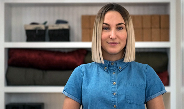 Eine junge Frau mit glatten blondgefärbten Haaren und einer Jeans-Bluse vor einem Regal mit Yoga-Utensilien