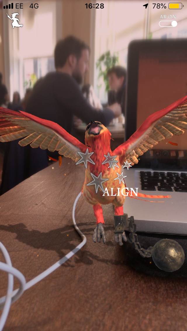 A tiny phoenix stretches its wings in Wizards Unite while in the background people sit in a cafe, going about their day.