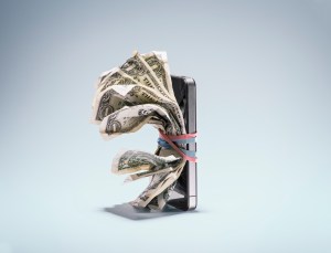 Five wrinkled dollar bills wrapped by a rubber band to the front of an iPhone.