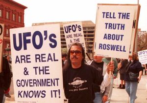 A protester in a black t-shirt holds signs reading "UFOs are real and the government knows it" and "Tell the truth about Roswell!"