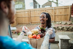 An over the shoulder shot of a young man holding a large bag of fruit and vegetables, his friend is smiling towards him.