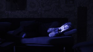 Eine Frau liegt zusammengekrümmt auf einem Sofa im Dunklen und hält sich schützend die Hände vors Gesicht. Sie sieht aus, als hätte sie Angst.