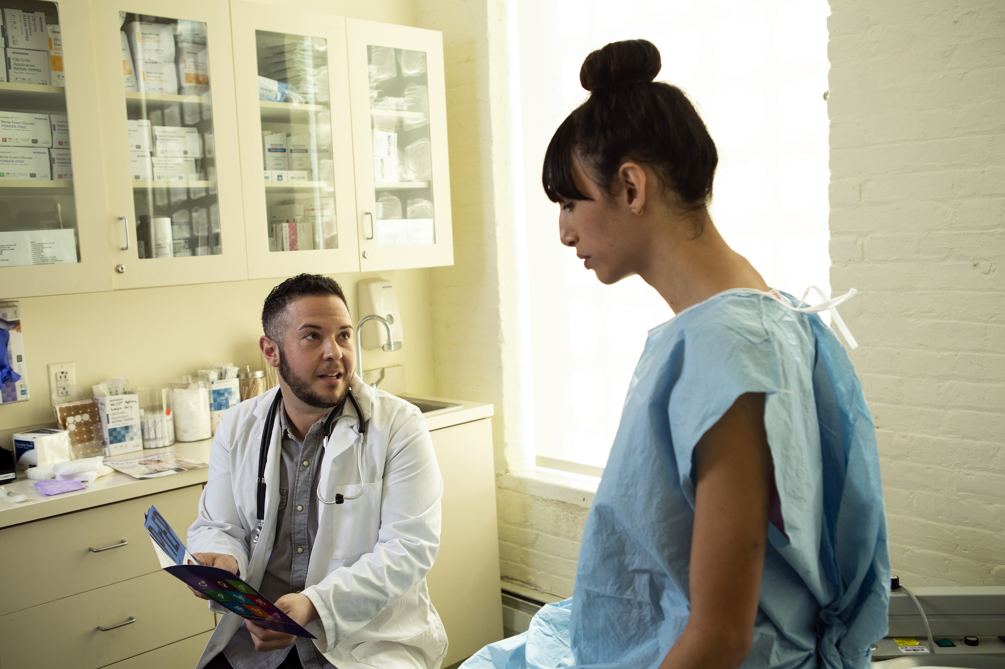 A-transgender-woman-in-a-hospital-gown-speaking-to-her-doctor-a-transgender-man-in-an-exam-room