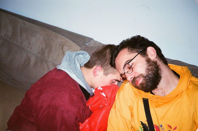 Two guys sleeping on a couch and leaning on each other.