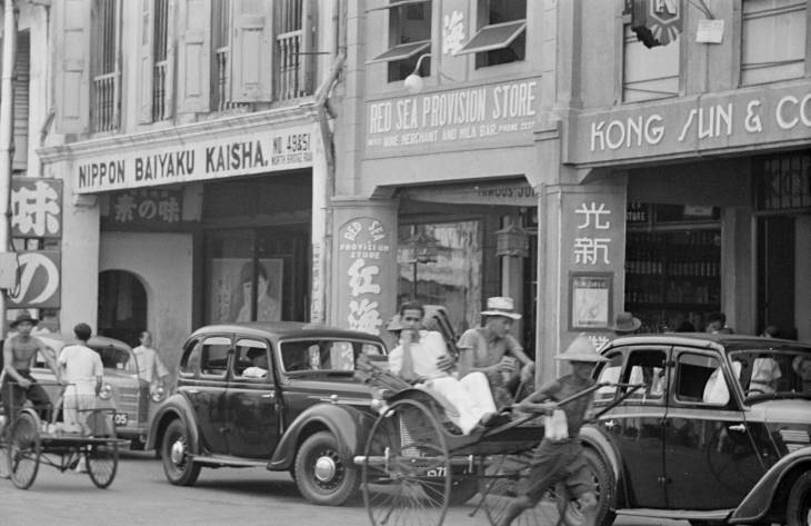rickshaw puller and cars driving in front of shops on a street in 1940s singapore
