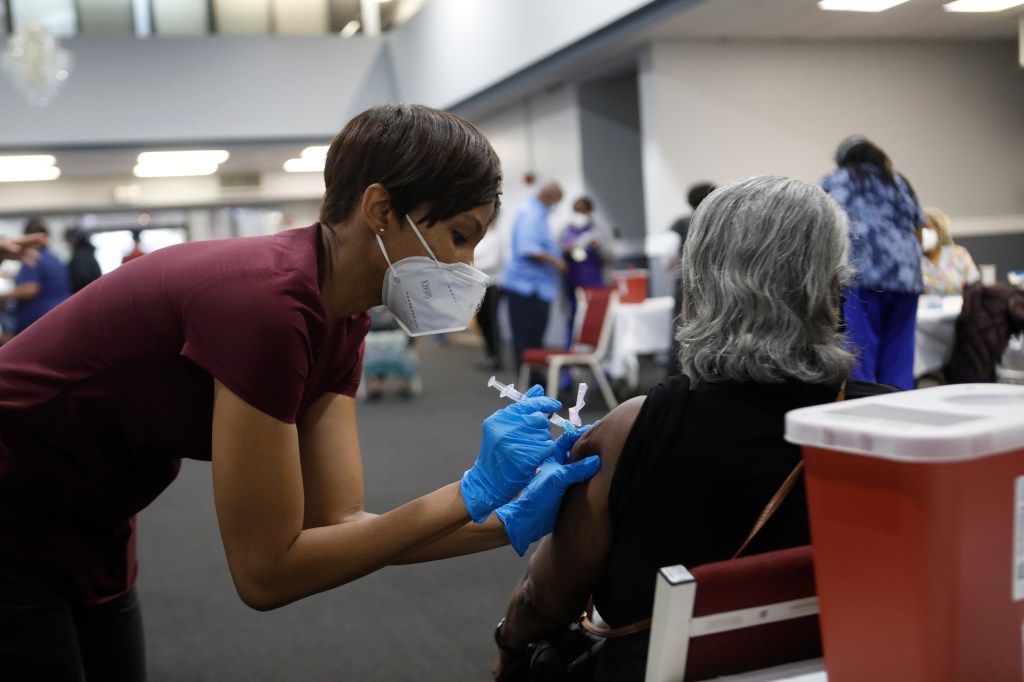 A woman with short hair administers a COVID-19 vaccine into the arm of a Black patient