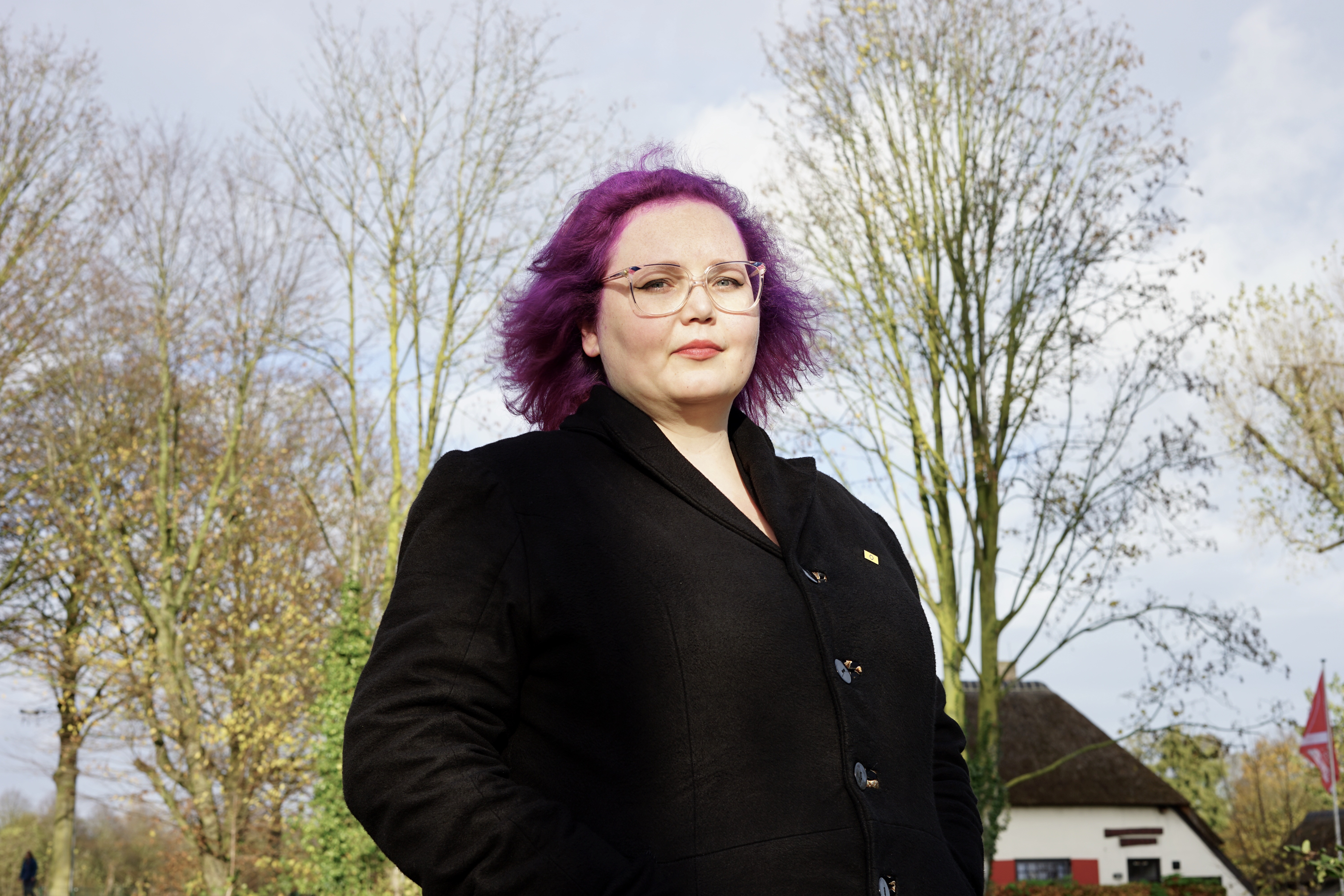 Annelies – woman with a purple bob and glasses standing outside in a residential area.