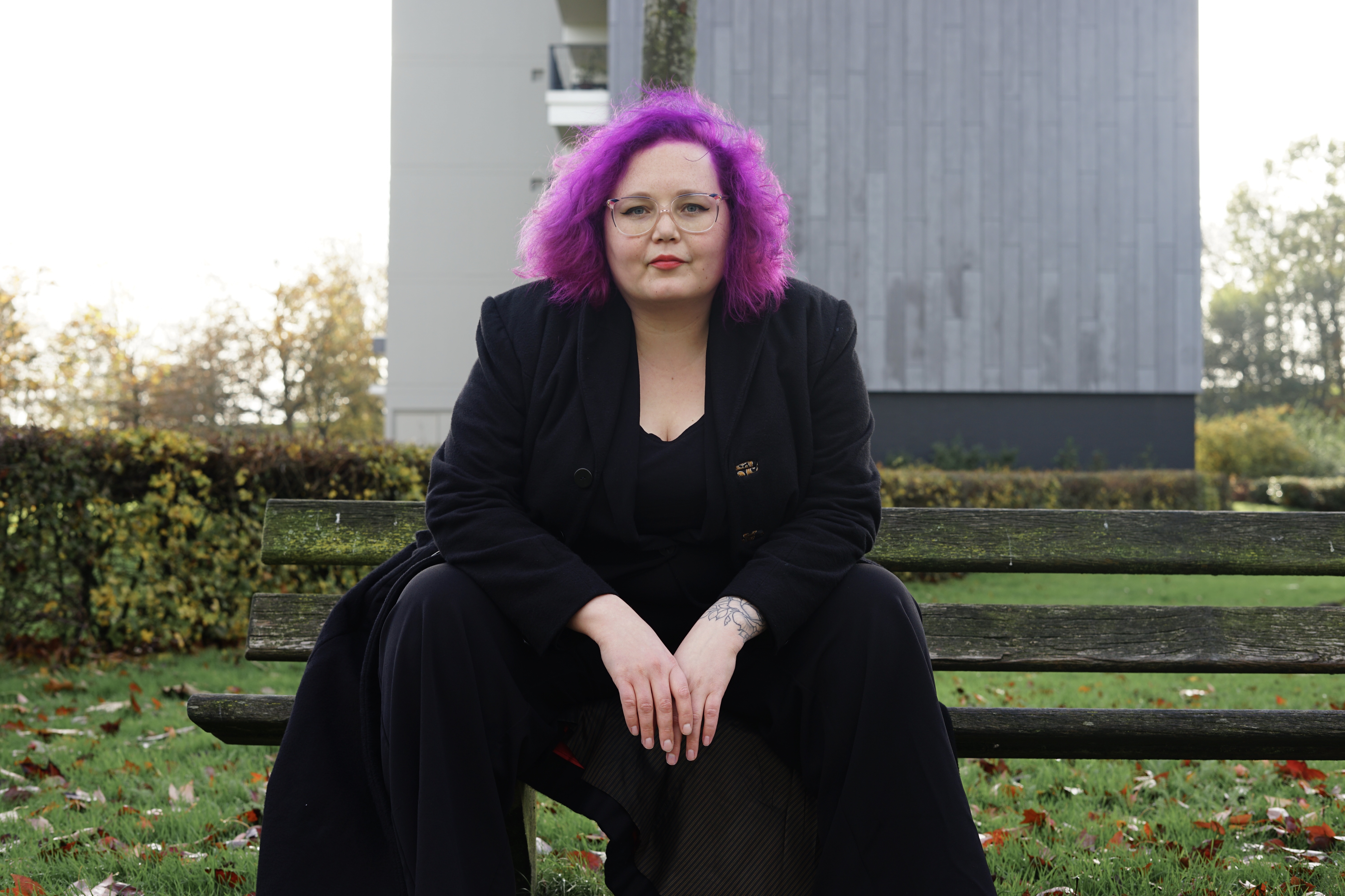 Annelies - woman with purple hair wearing a black outfit and sitting on a bench.