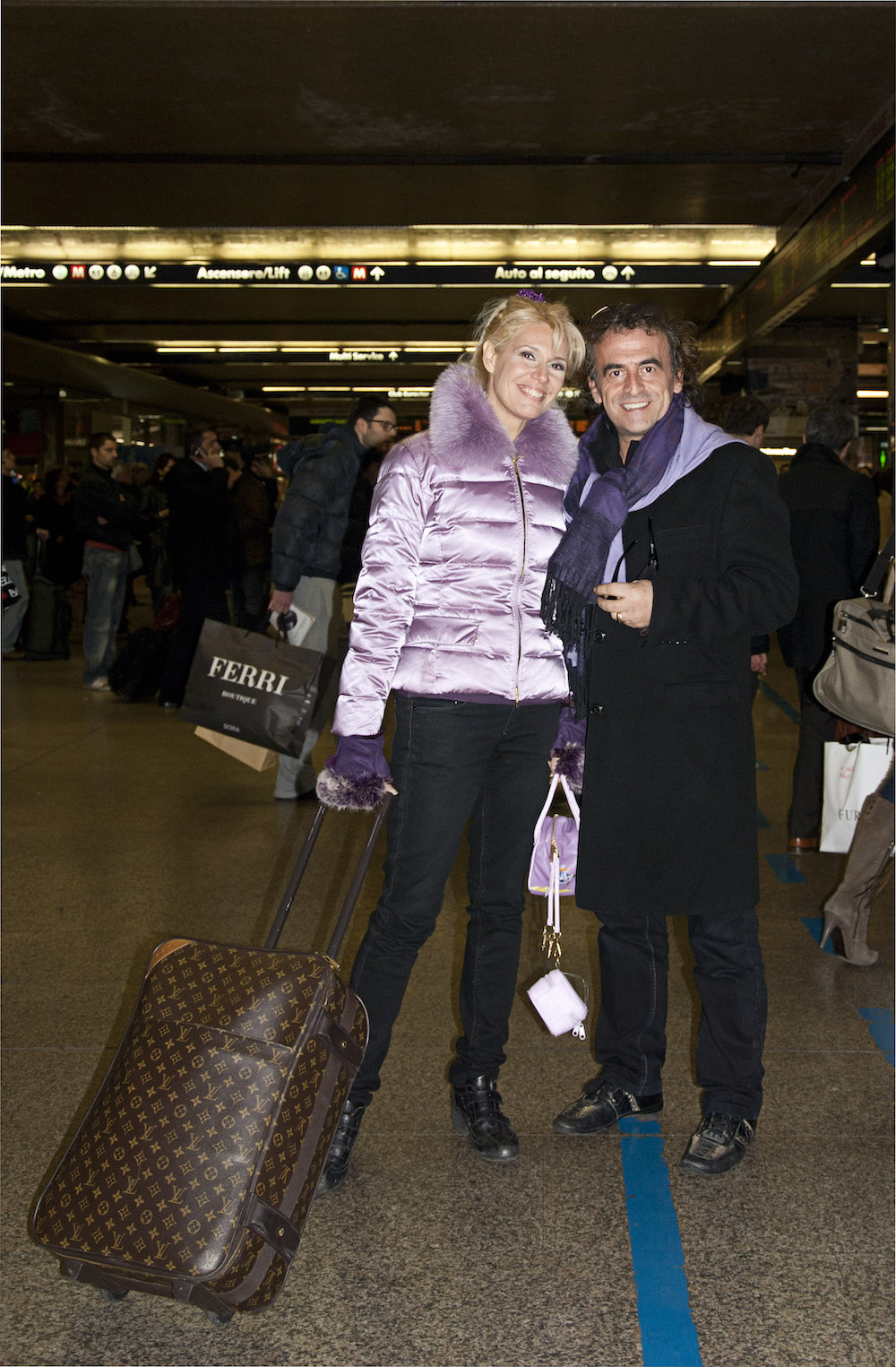 Eine blonde Frau mit Louis-Vuitton-Koffer und ein Mann in Trenchcoat posieren im Hauptbahnhof Roms für ein Foto