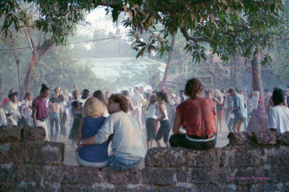 Menschen sitzen auf einer alten mauer, dahinter tanzen welche auf einem großen Platz zwischen Bäumen