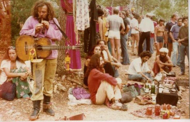 Ein Mann mit lange Haaren und Hippiekleidung spielt Flöte und hat eine Gitarre umgehangen, drumherum sitzen und stehen Leute