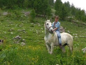 Roberta Colombero, margari – Roberta on her white horse in the middle of an Alpine meadow. She’s tall, thin with long dirty blonde hair in a ponytail and bangs.