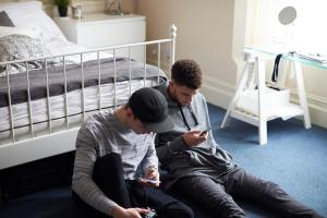 Two Male Students In Bedroom Check Messages On Mobile Phones