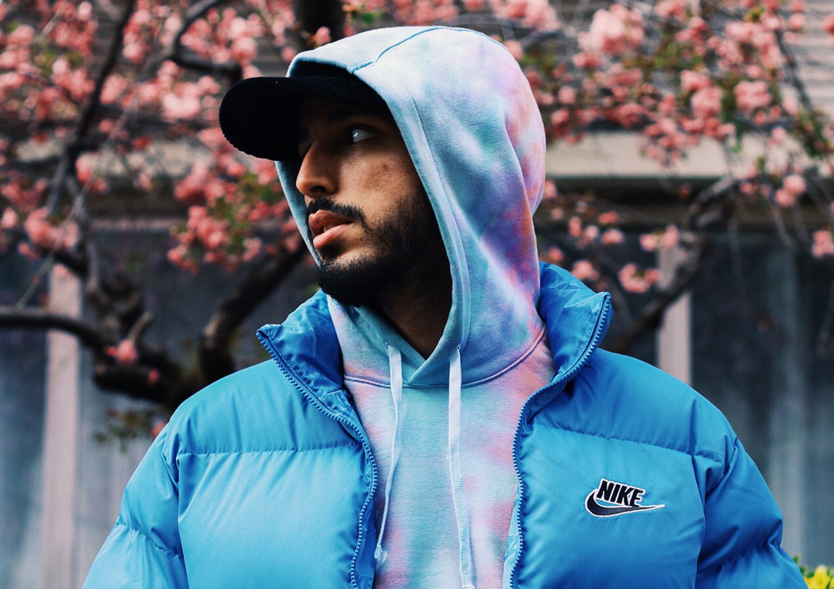 Tawsen – young man wearing a blue puffer jacket, a pastel tie-dye sweatshirt pulled over a baseball cap. Tawsen has a beard and is looking to the side as he poses in front of a tree in bloom.
