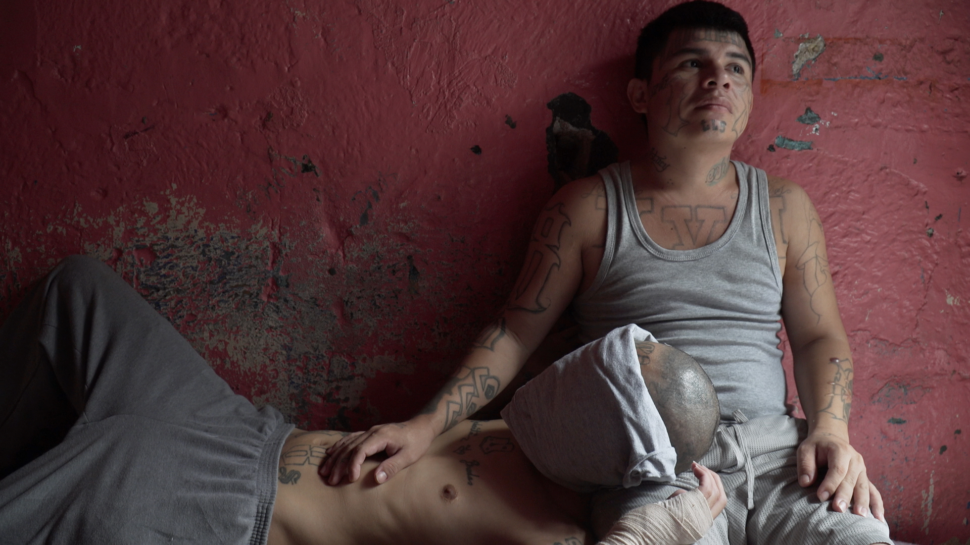 Zwei junge Männer mit vielen Gang-Tattoos sitzen vor einer roten Wand, einer hat seinen Kopf in den Schoß des anderen gelegt