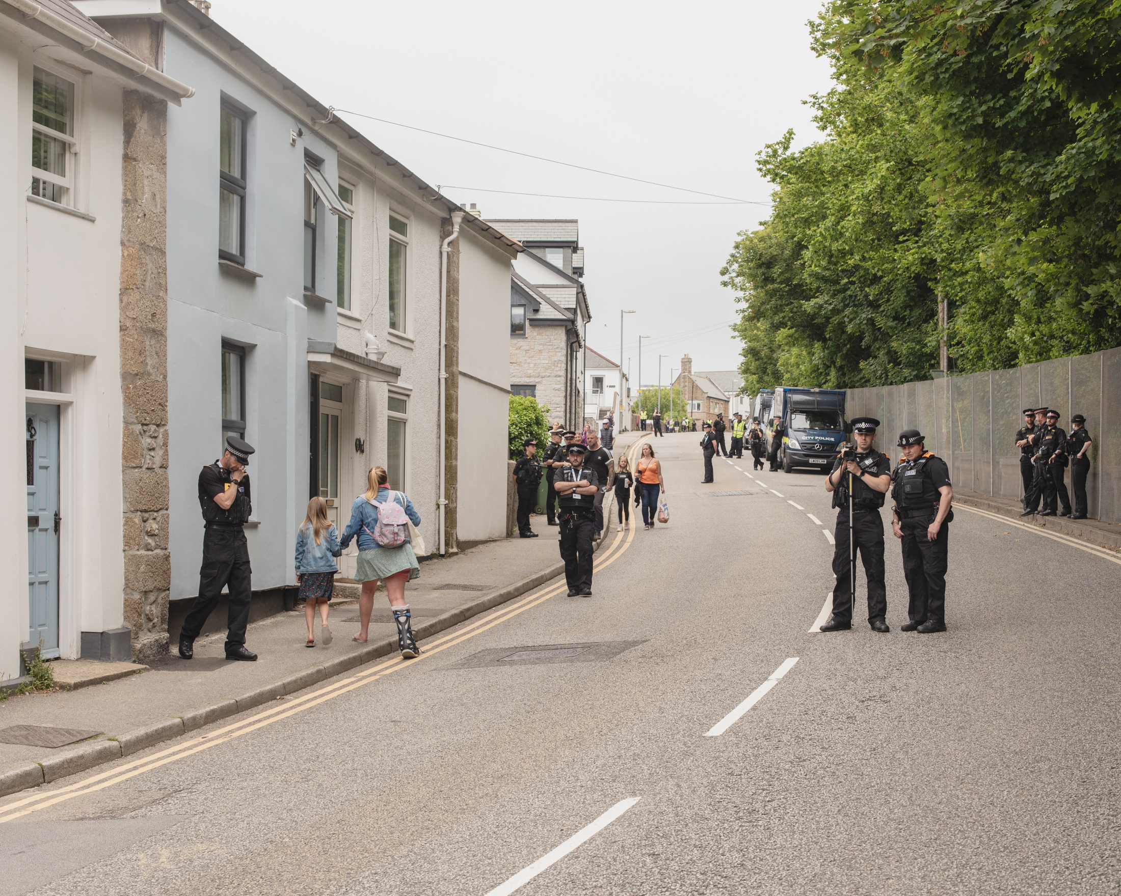 A heavy police presence outside of the steel wall erected around Carbis Bay, the site of the G7 summit. Locals walk among them, going about their day.