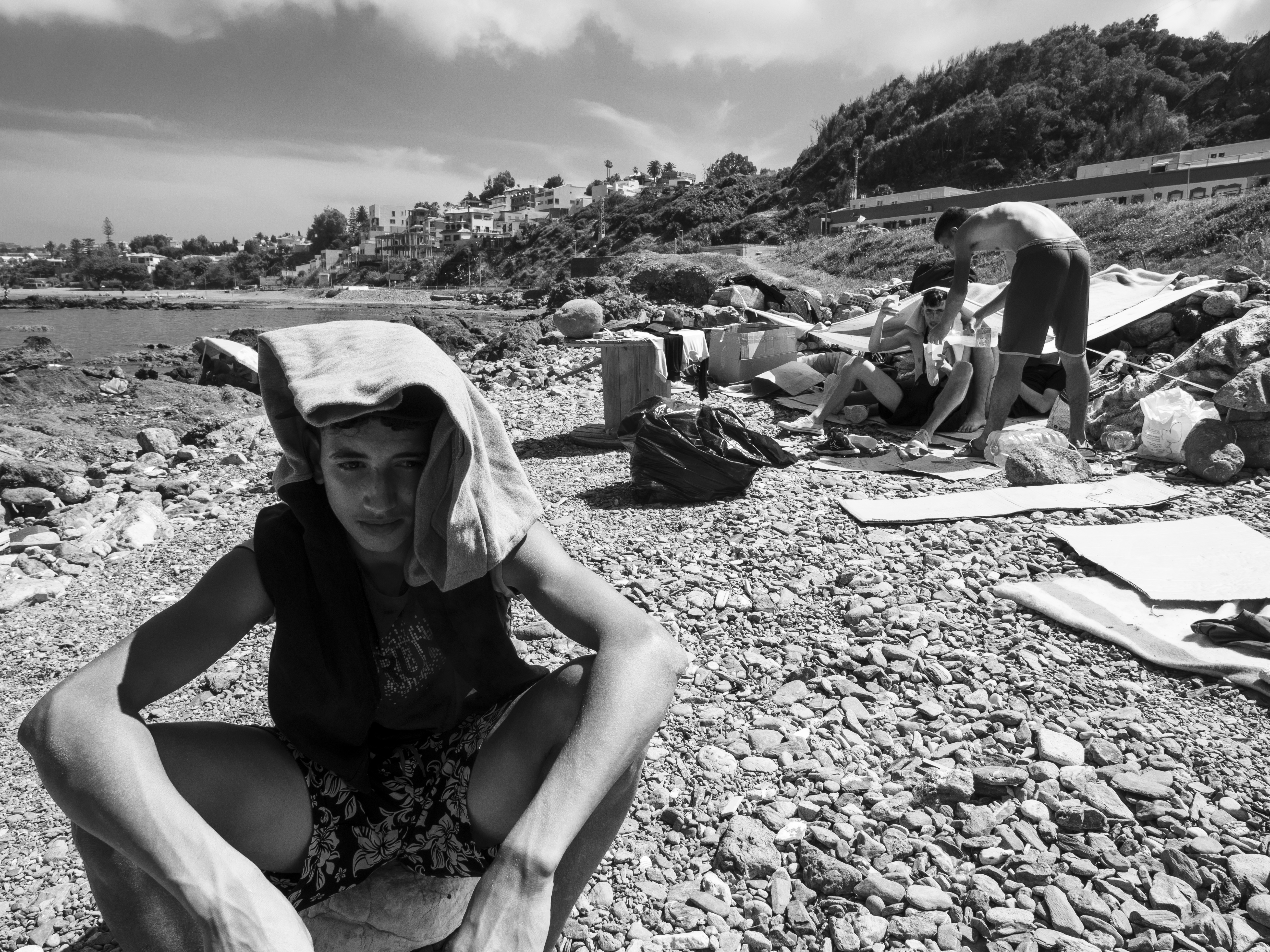 Hervé Lequeux, Ceuta – Drie jongeren zitten op een stenen strand omringd door kartonnen dozen. De jongen op de voorgrond beschermd zijn hoofd met een kledingstuk tegen de zon