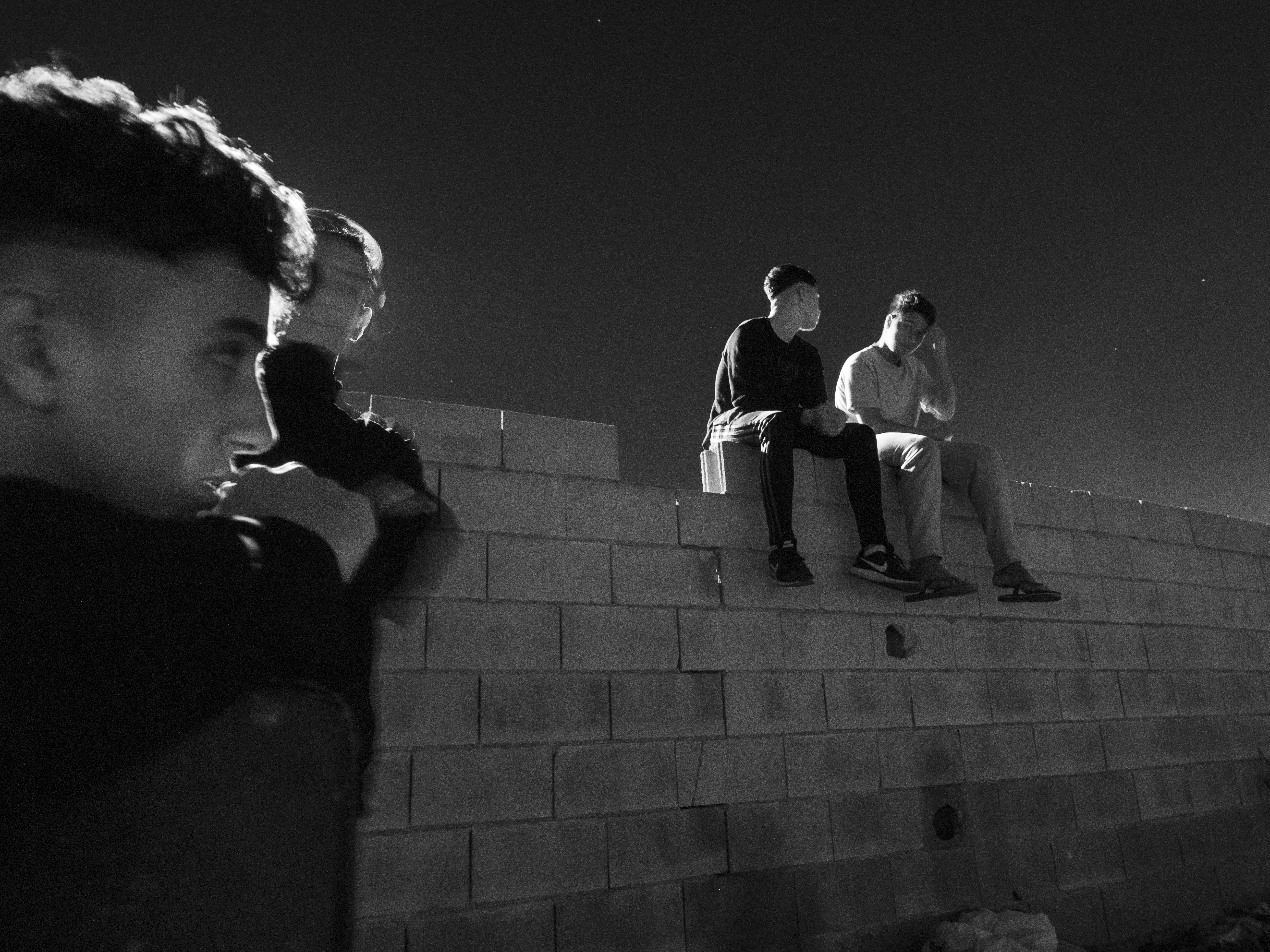 Hervé Lequeux, Ceuta – Two teens talking and sitting on a wall. Off to the side, another teen is looking at them with a fourth standing behind him.