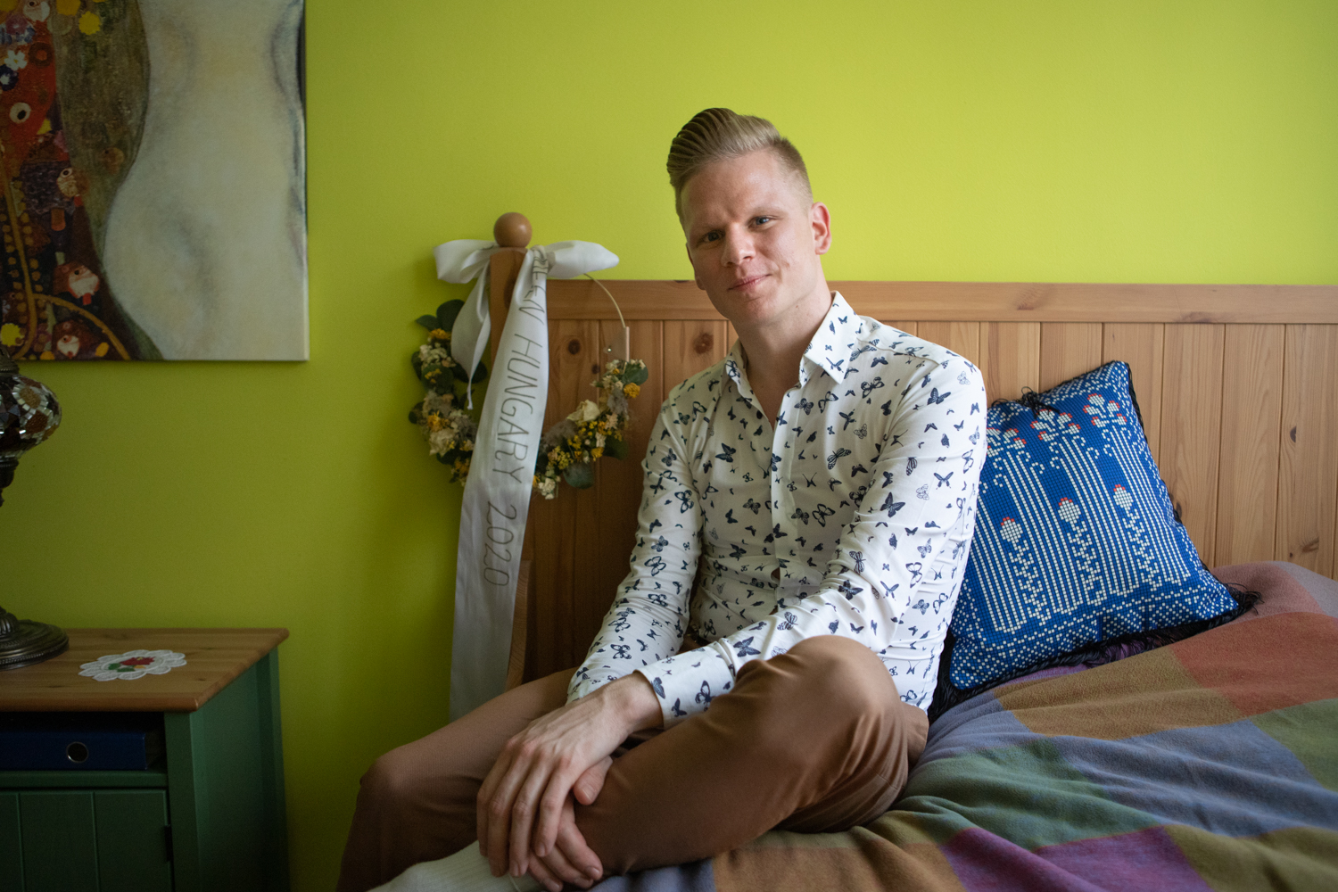 Ein junger Mann in hellem Hemd und brauner Hose sitzt bei sich zu Hause auf dem Bett und blickt in die Kamera