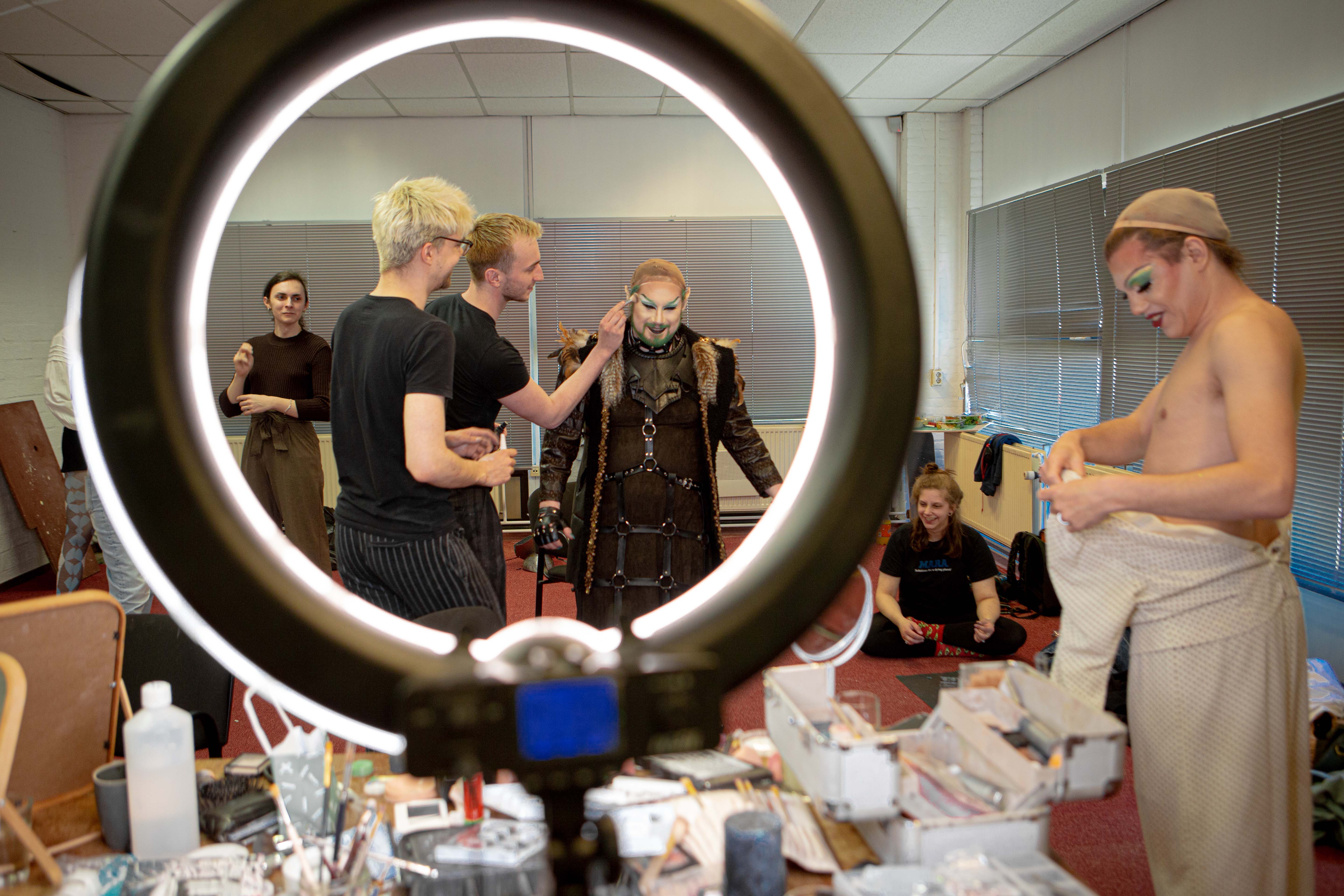 Queer Arcana – seven people getting dressed and putting makeup on in a crammed room. In the foreground there's a big ring light