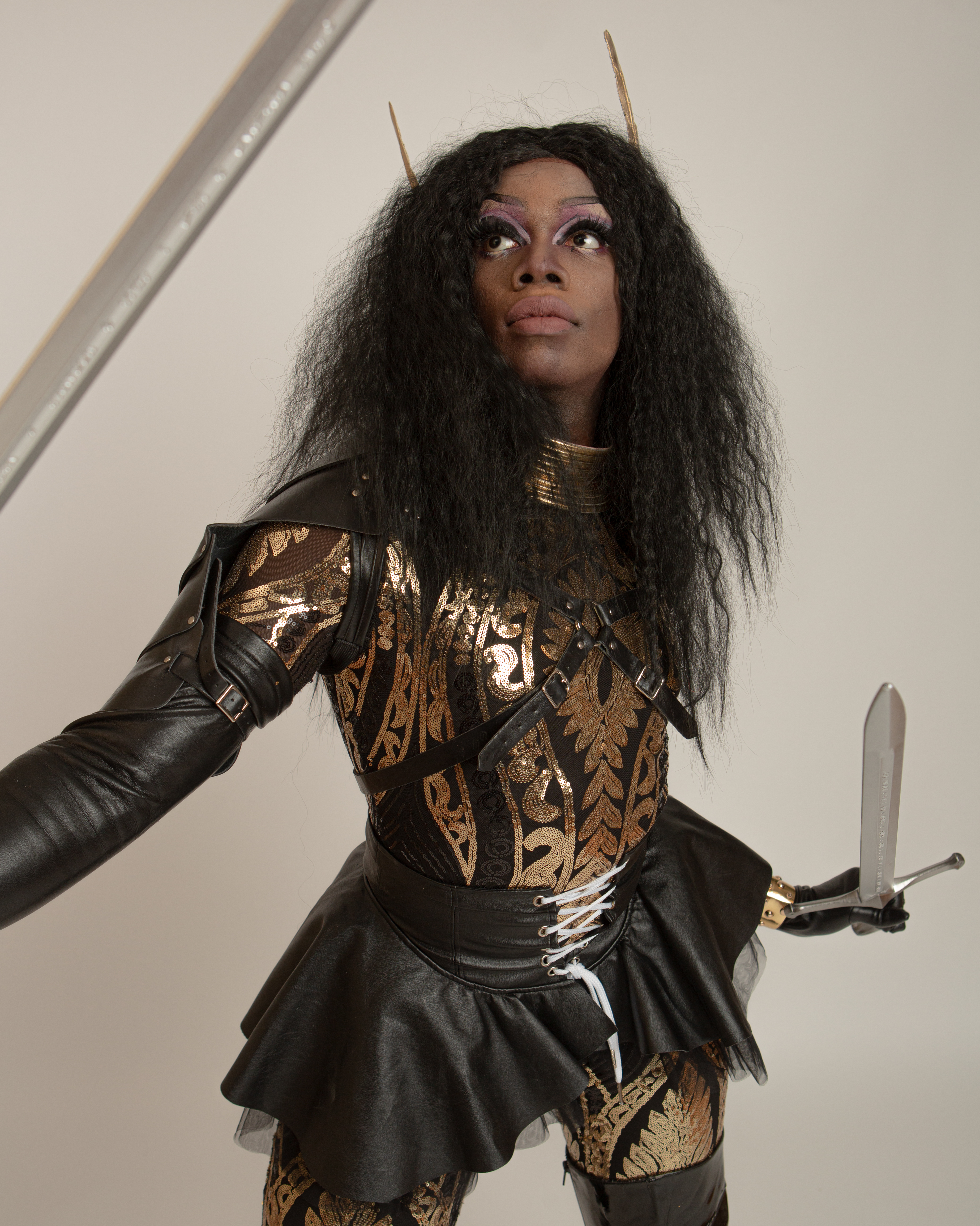 John Togba, Queer Arcana – female character wearing a black and gold patterned bodysuit, a black leather tutu skirt and golden horns, brandishing a sword.