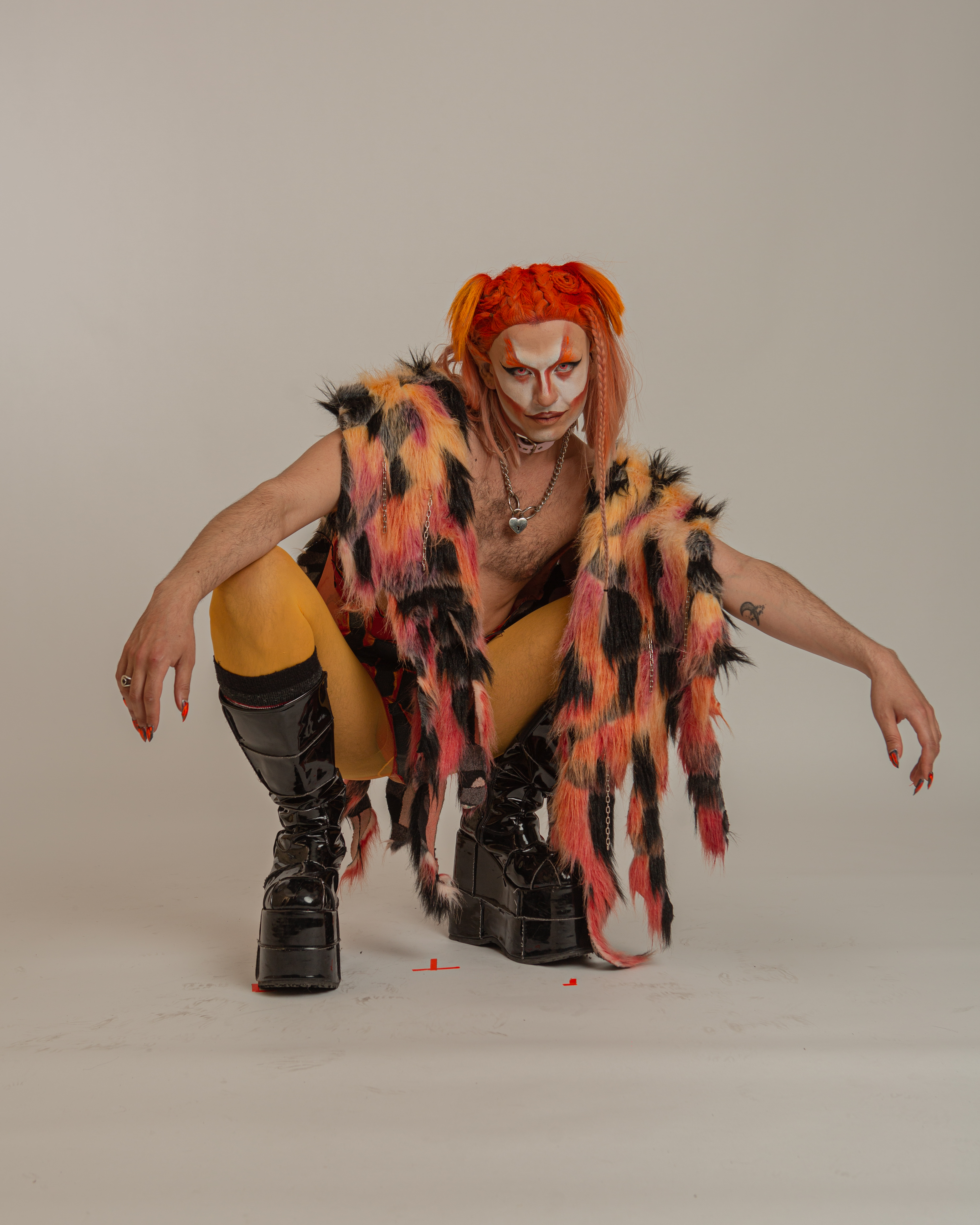 Alain Chaney, Sasa Hara, Queer Arcana – bare-chested character wearing orange tights, platform combat boots, a red, yellow and black fur shawl and a braided orange wig.
