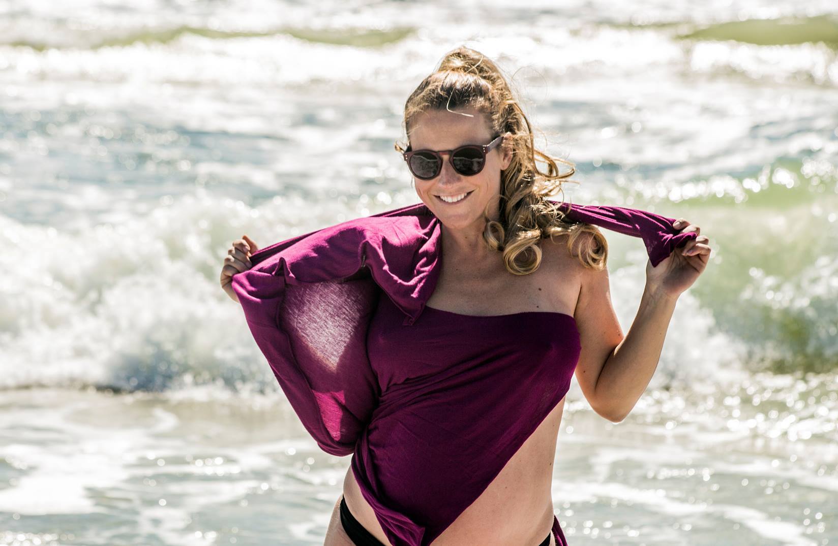 Eine lächelnde Frau mit blonden Haaren, einem violetten Top und einer Sonnenbrille posiert am Strand