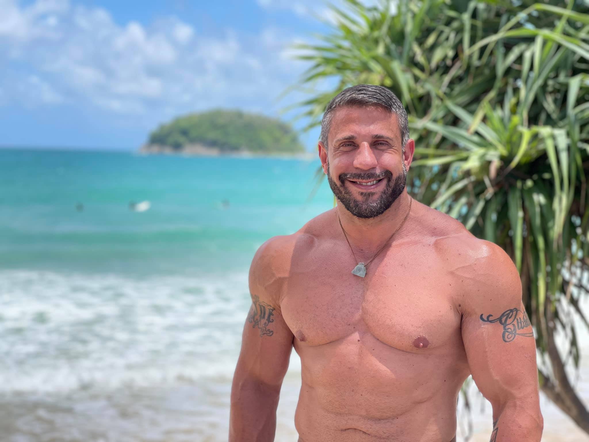 Ein muskulöser Mann mit Bart und kurzen grauen Haaren steht oberkörperfrei an einem Strand mit türkisblauem Wasser im Hintergrund