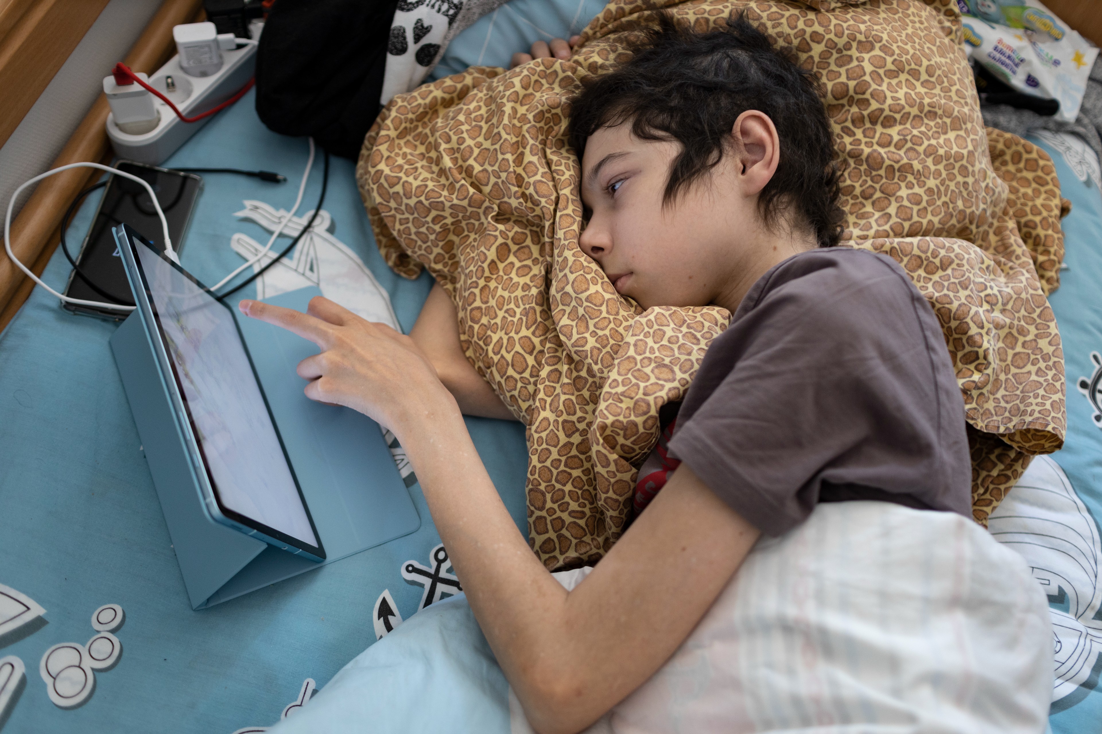 Ein dünner Junge mit dunklen Haaren liegt im Bett und bedient ein Tablet