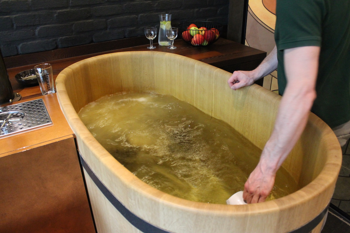 Bart & Barley, beer spa in Belgio – Una vasca di legno contenente un liquido giallastro frizzante e una mano che aggiunge ingredienti