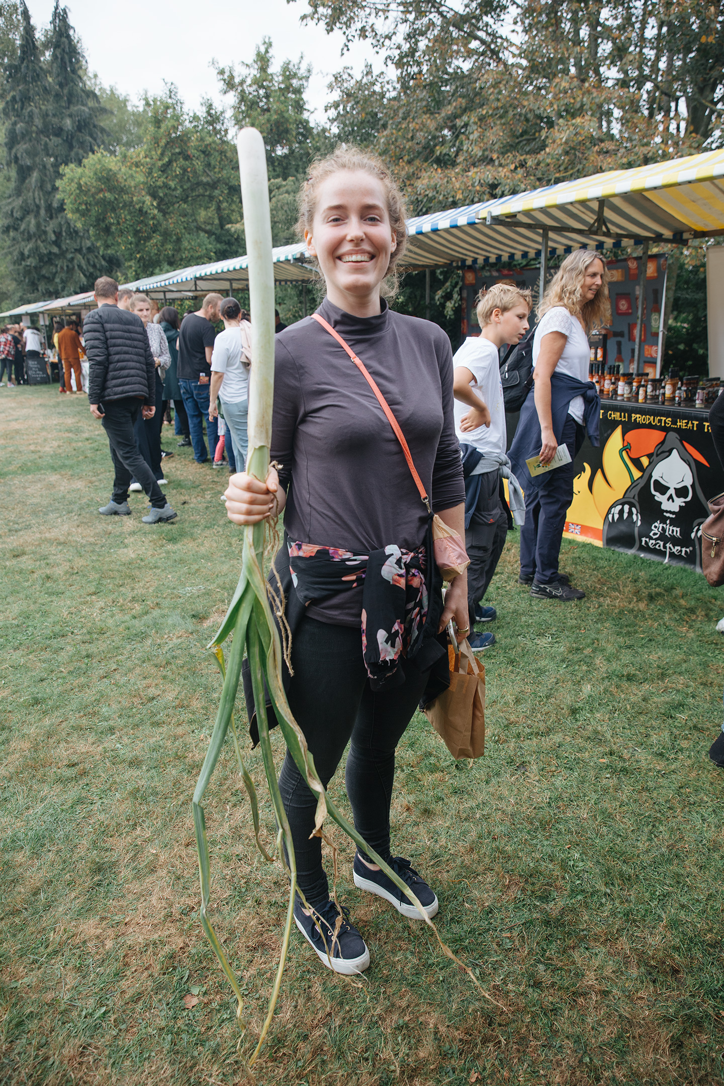 Raymond van Mil, Netherlands, pumpkins - woman dressed in black holding a large, thin, green leek in front of a row of market stalls.