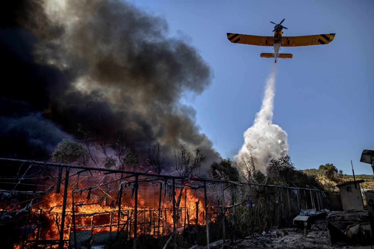 In “Fire Destroys the Moria Camp 03” by Jacob Ehrbahn from Denmark, a plane drops water over Moria refugee camp as it burns on the 10th of September 2020 . Photo: Jacob Ehrbahn