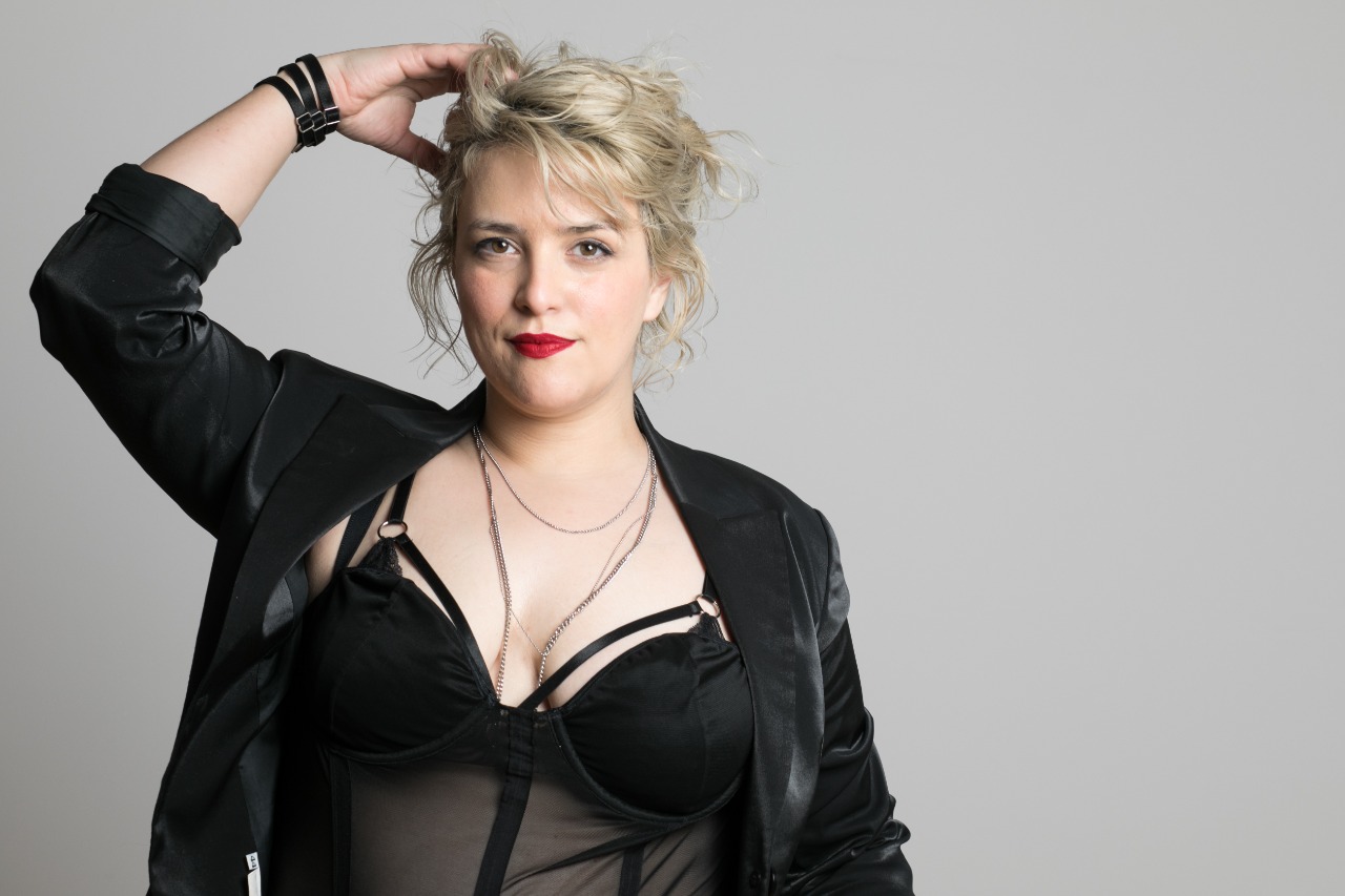 Mainstream porn, sex education - Photo of a blonde woman wearing a black top stood in front of a grey background.