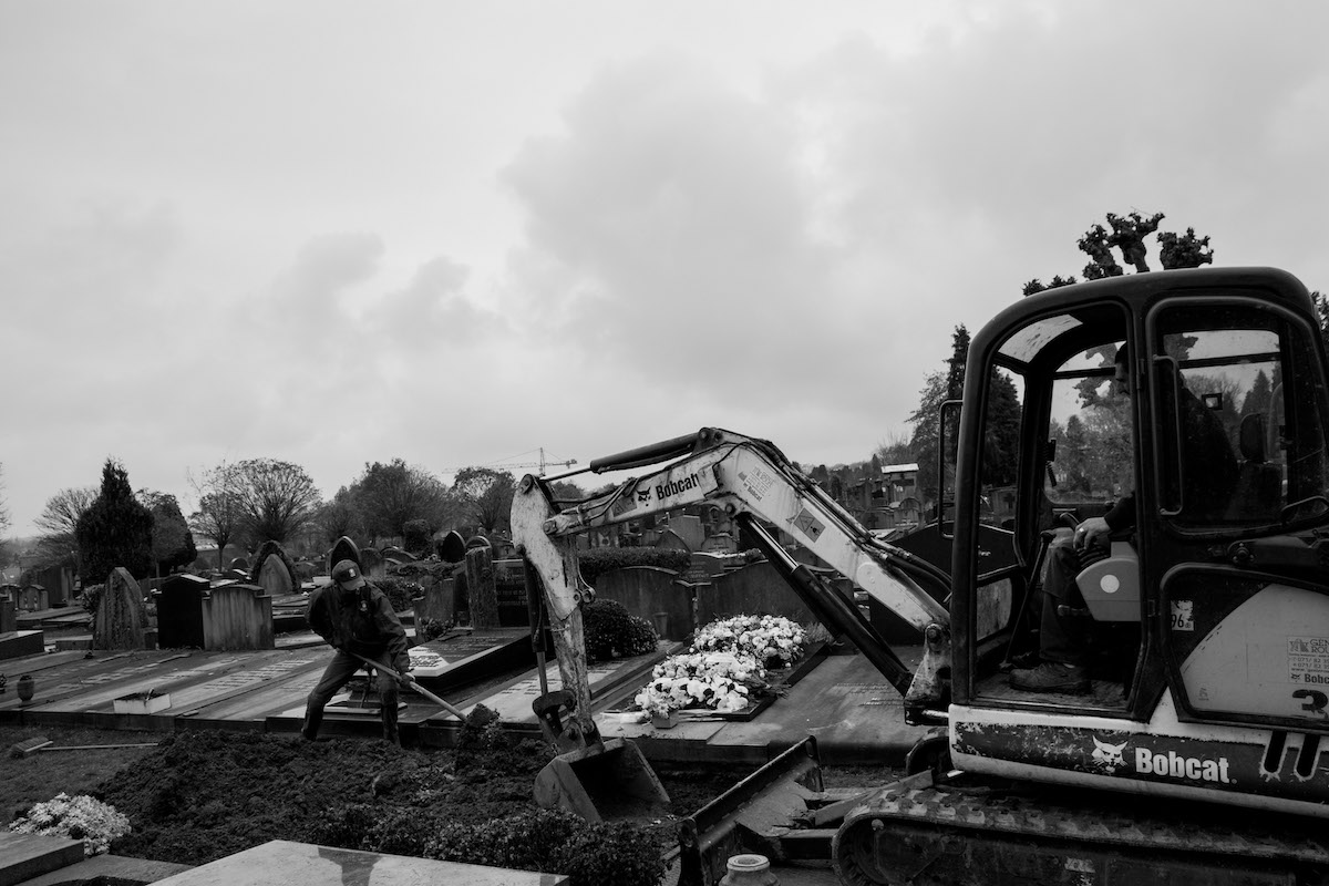 Jacques Vermeer, Patrick, cemetery - Black and white photo of a man digging a grave with a shovel, to his right is a mechanical digger
