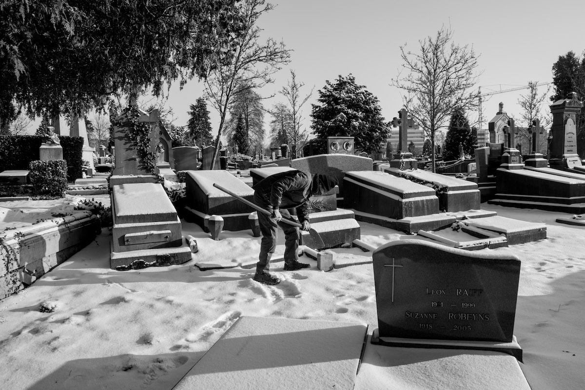 Jacques Vermeer, Patrick, cemetery - Black and white photo of a man with a shovel stood in the snow, surrounded by tombstones.