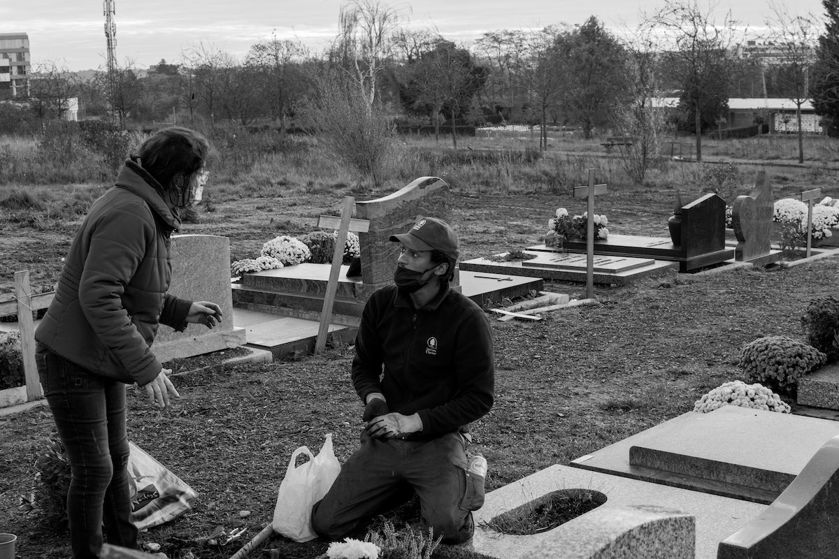 Jacques Vermeer, Patrick, cemetery - Black and white photo of a woman in a cemetery talking to a kneeling man in a baseball cap and face covering, next to a tomb.