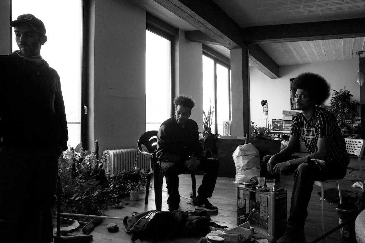 Jacques Vermeer, Patrick, cemetery - Black and white photo of three men sat in an apartment, all looking at something out of shot.