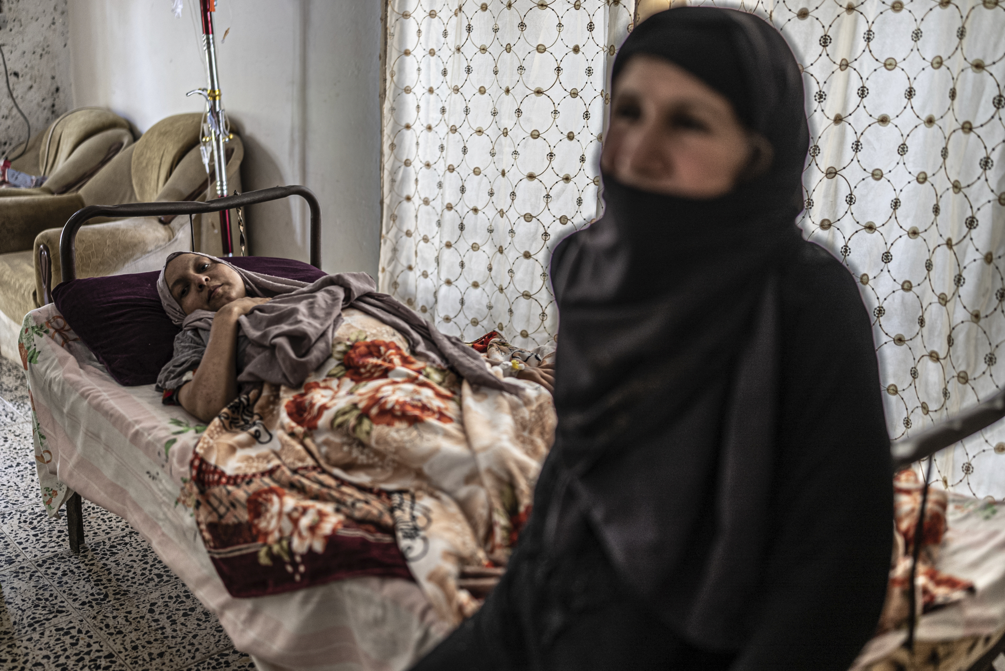 Climate crisis, Iraq - Background, sick woman lying on hospital bed attached to a drip, foreground woman in traditional black dress.