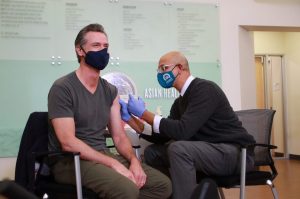 Gavin Newsom in a black mask receives a COVID booster shot from a doctor in a dark blue sweater and light blue mask.
