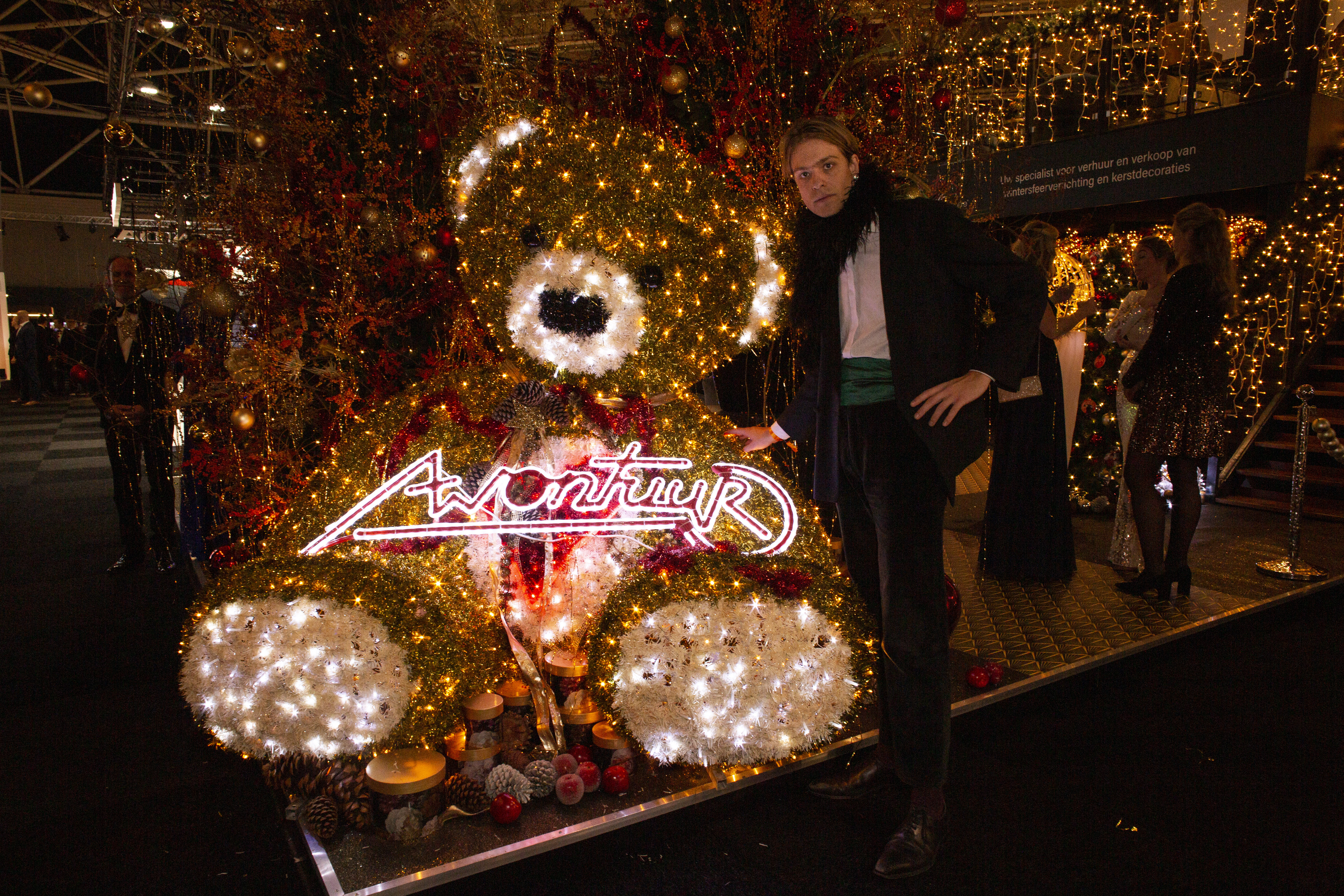 Masters expo, wealth, roaring twenties - Man in tuxedo stands next to a human-sized gold teddy bear.