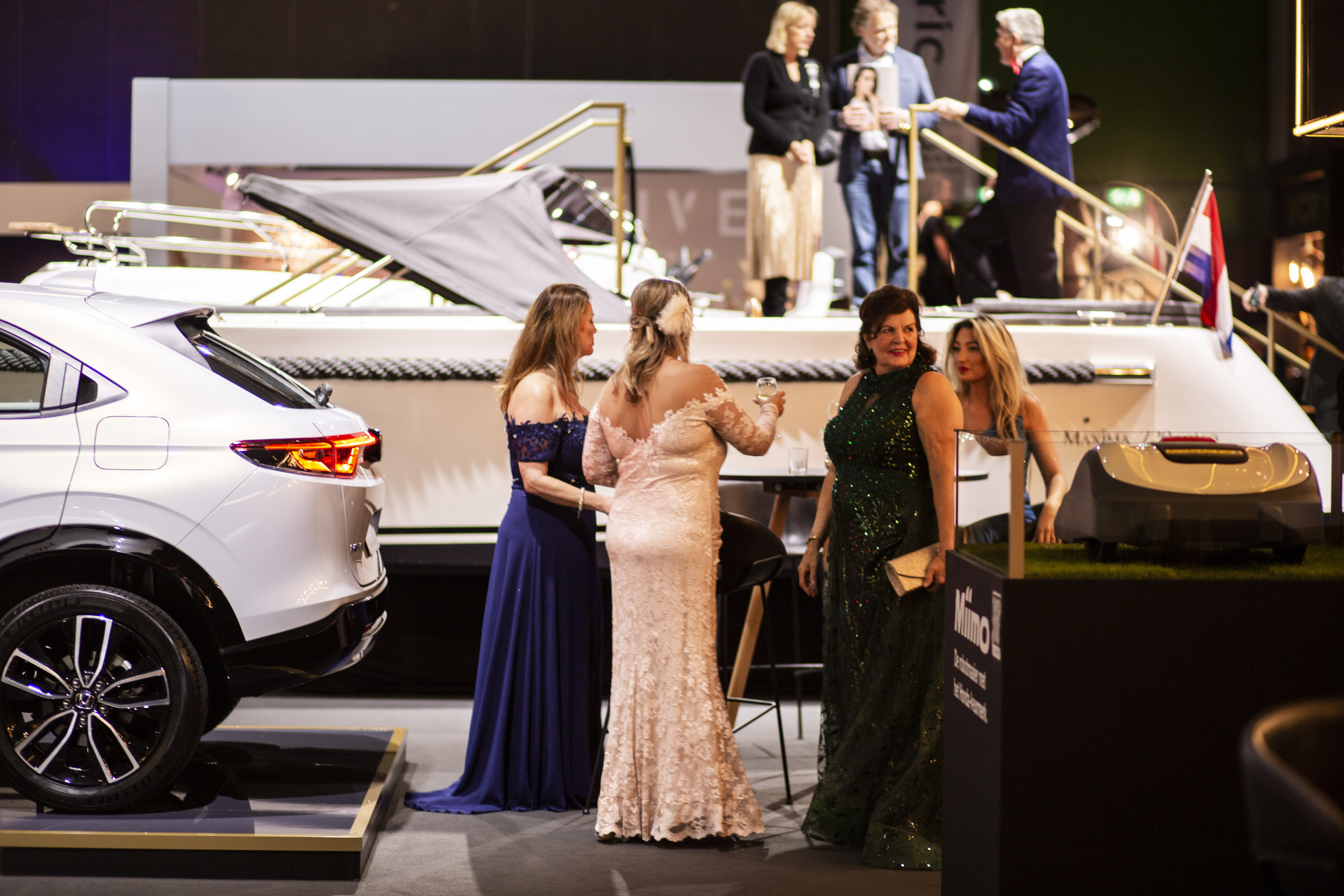 Masters expo, wealth, roaring twenties - Photograph of four women wearing dresses stood in front of a luxury yacht and a white car.