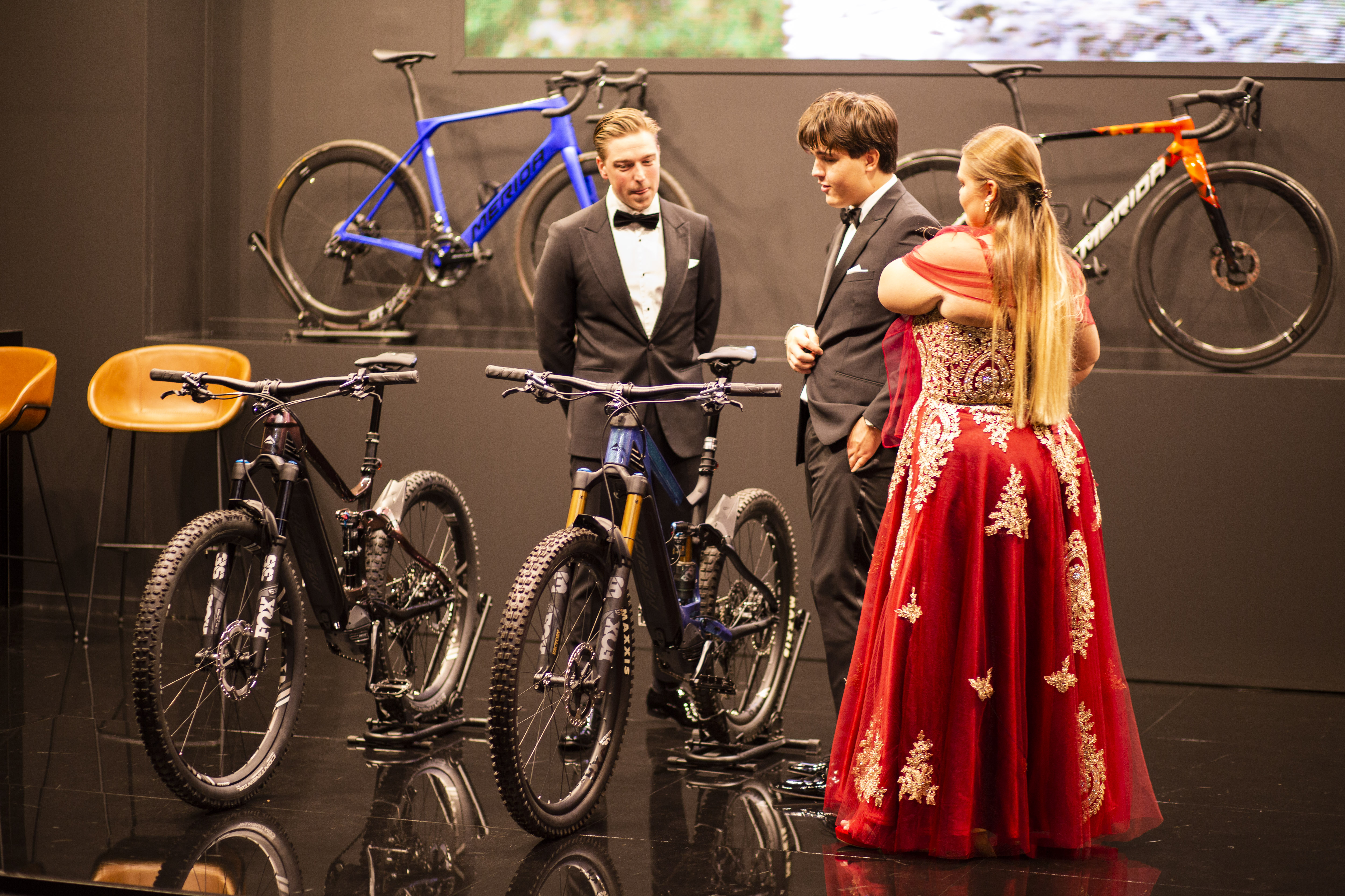 Masters expo, wealth, roaring twenties - Two men in tuxedos and a woman in a red dress look at a collection of four bikes.