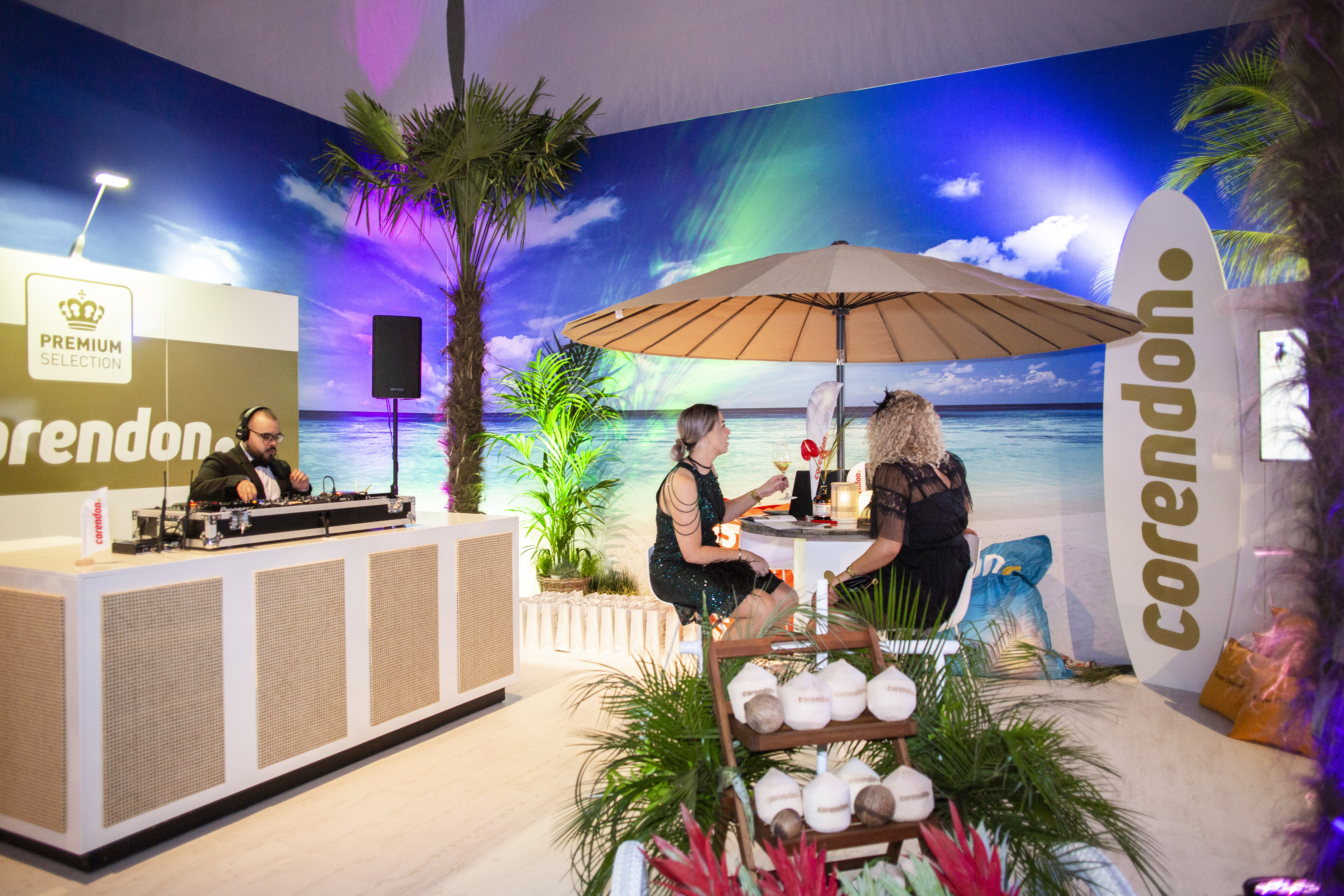 Masters expo, wealth, roaring twenties - A DJ in a tuxedo plays music to two women sitting under a parasol in a room designed to look like a sandy beach.