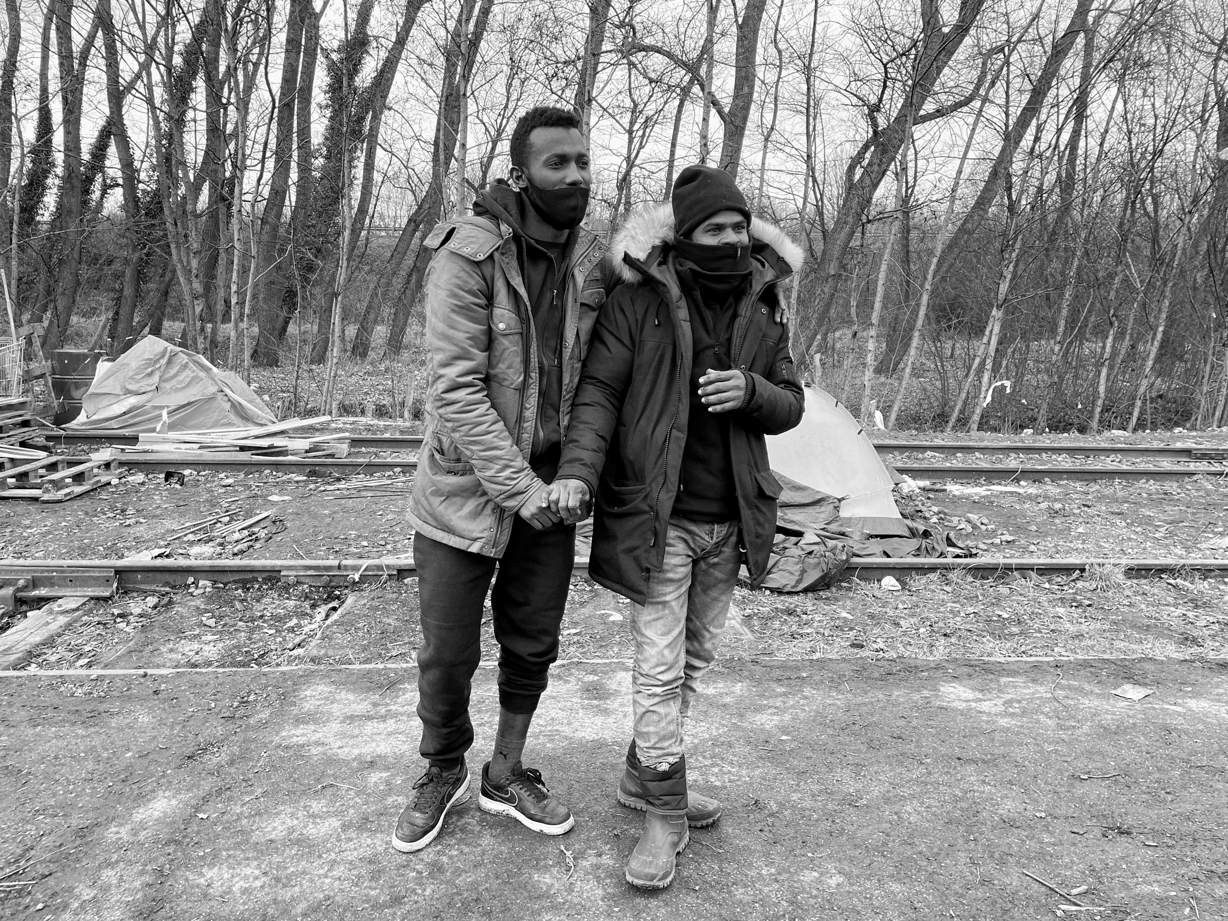 Calais, refugee camps - Black and white photo of two men wearing coats, holding hands in front of a line of thin trees.