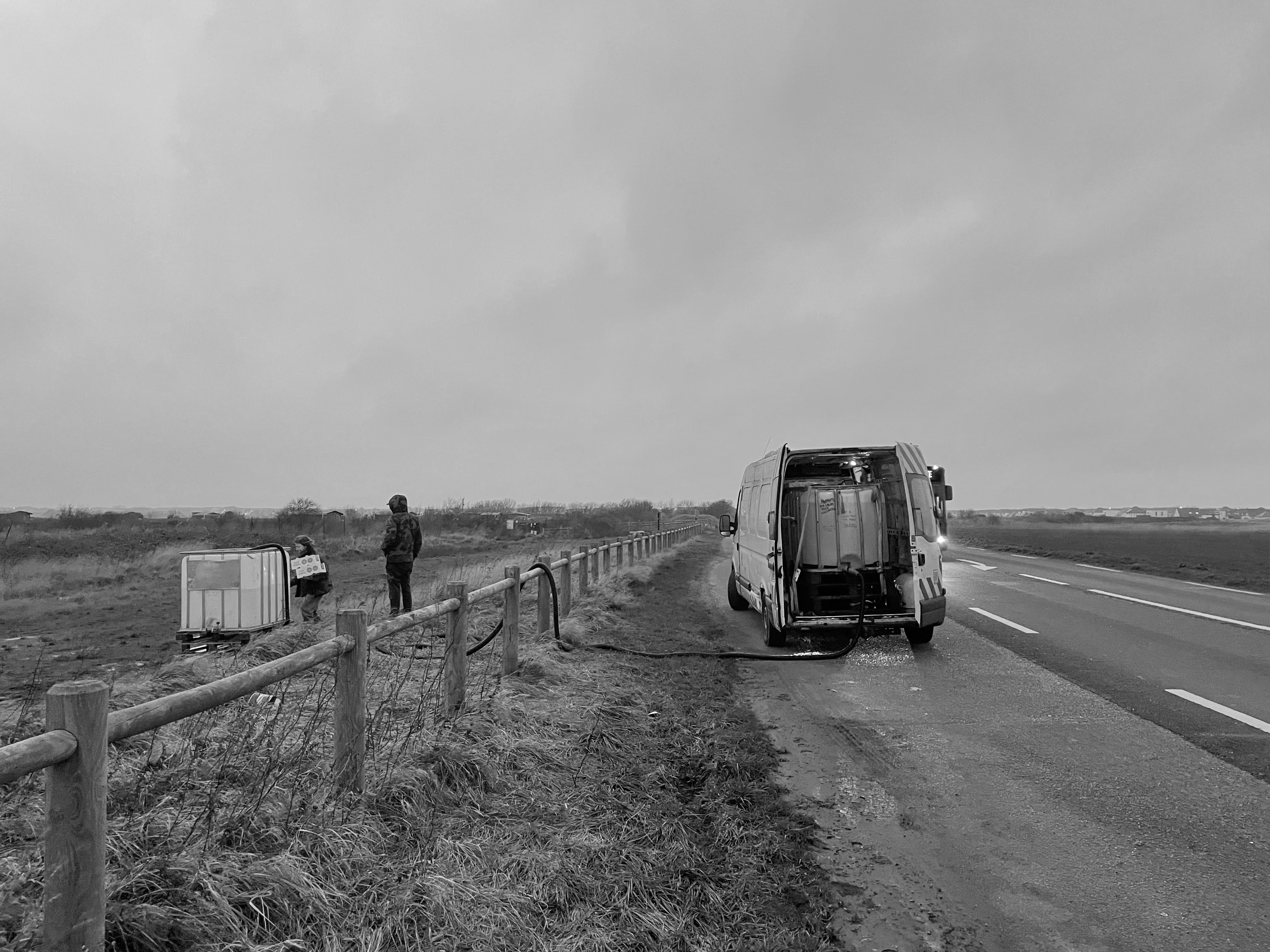 Calais, refugee camps - Black and white photo of a tank being filled with water in the back of a van on the side of an empty road.