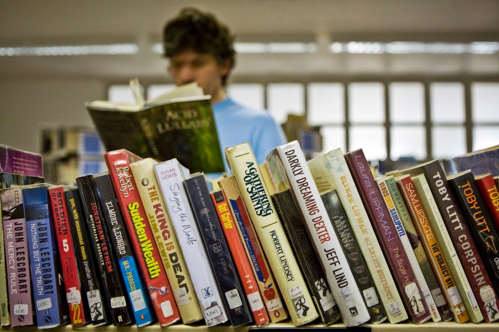 A row of books on a shelf at a prison library with a person reading out-of-focus behind